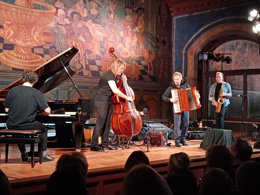 Vier Musiker treten auf der Bühne mit einem Flügel, einem Kontrabass, einem Akkordeon und einem Saxophon auf. Sie spielen vor einem großen, farbenfrohen Wandgemälde, während das Publikum ihnen zusieht. Der Raum ist warm beleuchtet und kunstvoll gestaltet.