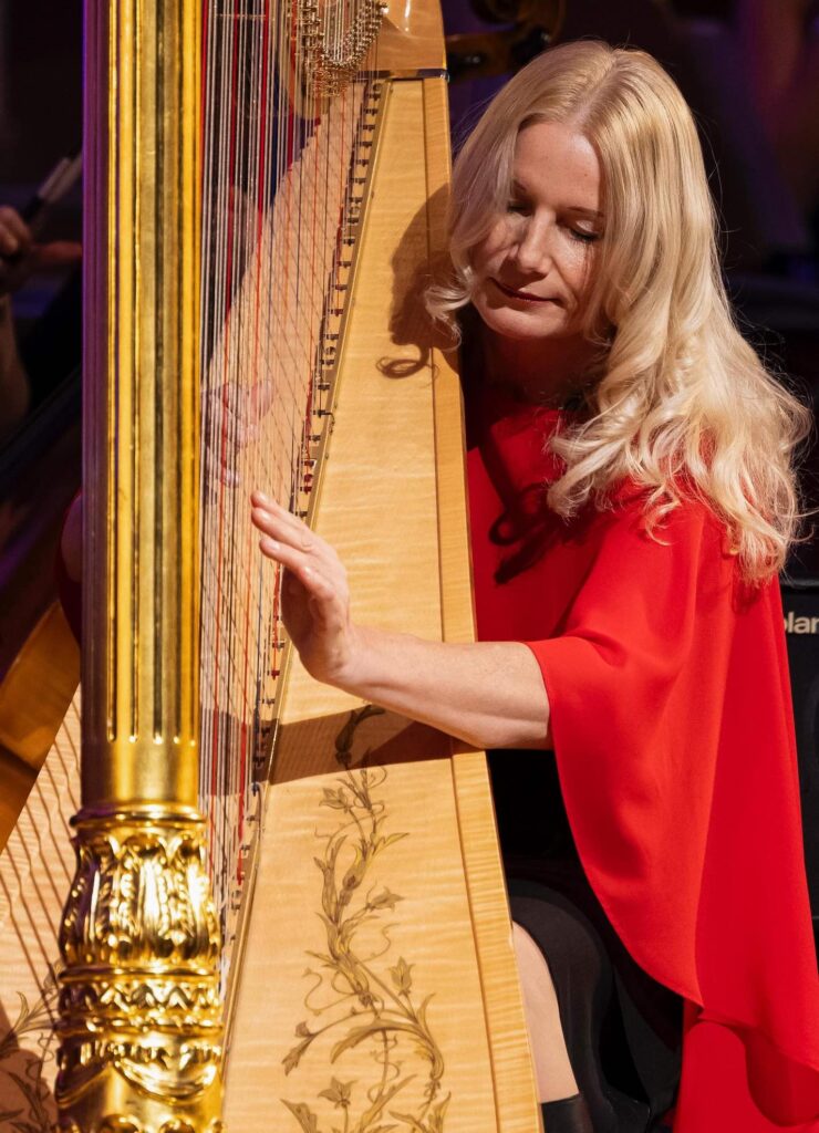 Eine Frau mit langen blonden Haaren in einem roten Kleid spielt auf einer goldenen Harfe und ist ganz in ihre Musik vertieft, während ihre Finger die Saiten zupfen.